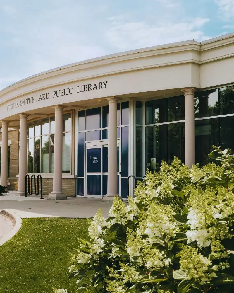 Exterior photo of the NOTL library. There are a series of white pillars along the facade of the building and a hydranga bush in full bloom in the foreground. The flowers are cream coloured.
