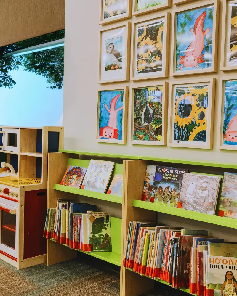 Image of a children's bookshelf, lime green, with picture books on a low shelf. The wall behind the shelf has a grid of children's paintings, featuring colourful flowers, penguins and pink axolotls. There is a large window to the left of the paintings with a bright blue sky and green tree branches.