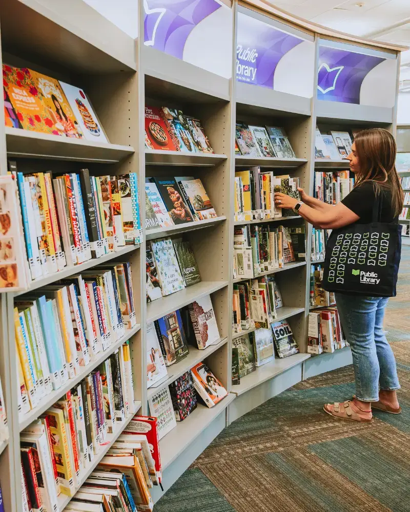 A large and colourful bookshelf spans the image. A young woman holding a navy blue Niagara-on-the-Lake tote bag is perusing the shelf. There is a purple toned sign above the shelf with the Niagara-on-the-Lake Public Library logo running across.